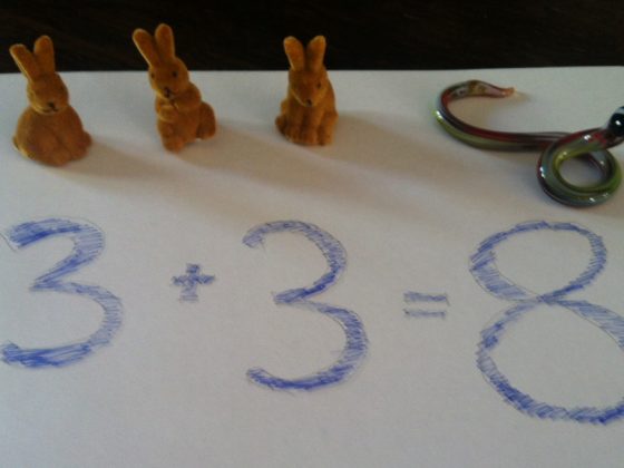 Zwei Deko-Osterhäschen sitzen auf einem Blatt Papier, auf dem mit blauer Farbe Zahlen aufgemalt sind in Form einer Gleichung 3+3=8.