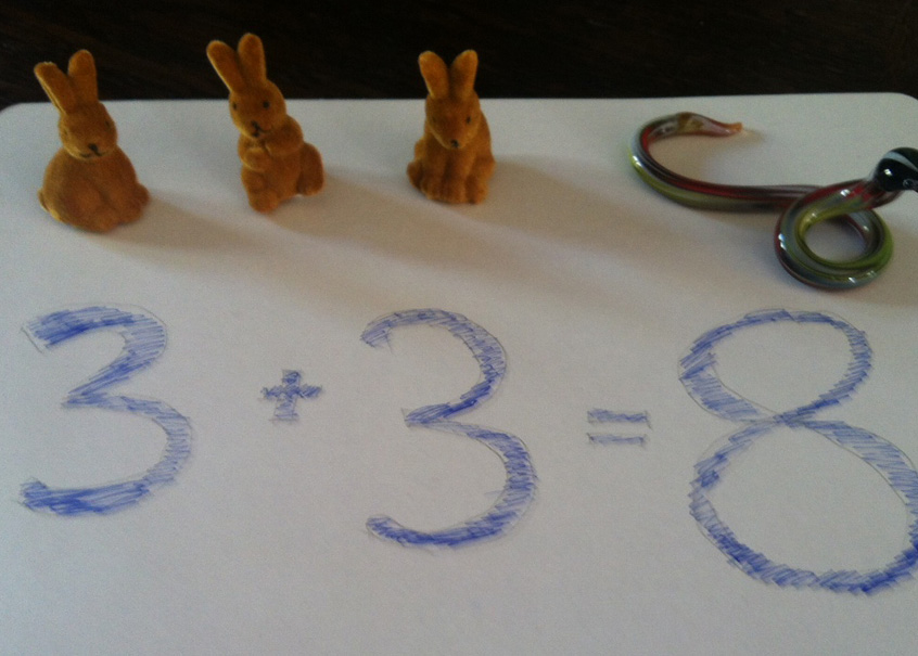 Zwei Deko-Osterhäschen sitzen auf einem Blatt Papier, auf dem mit blauer Farbe Zahlen aufgemalt sind in Form einer Gleichung 3+3=8.
