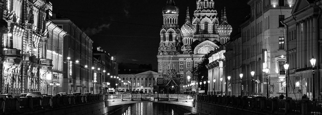 Die Basilius-Kathedrale in Moskau bei Nacht. Sie ist hell angestrahlt, ebenso wie die prachtvollen Häuserfassaden entlang eines Kanals, der auf die Kirche zuläuft.