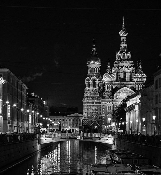 Die Basilius-Kathedrale in Moskau bei Nacht. Sie ist hell angestrahlt, ebenso wie die prachtvollen Häuserfassaden entlang eines Kanals, der auf die Kirche zuläuft.
