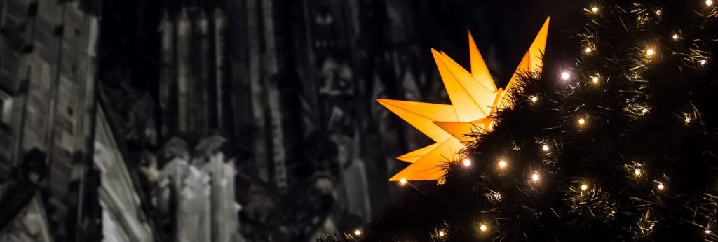 Vor einer Teilansicht des Eingangsportals des Kölner Domes steht ein mit einem großen, leuchtenden Weihnachtsstern und elektrischen Kerzen geschmückter Christbaum.