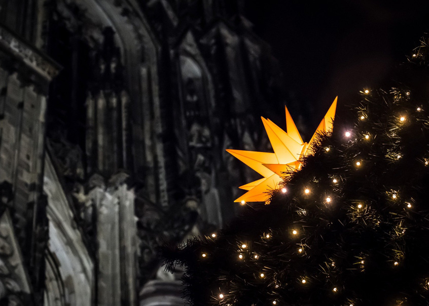 Vor einer Teilansicht des Eingangsportals des Kölner Domes steht ein mit einem großen, leuchtenden Weihnachtsstern und elektrischen Kerzen geschmückter Christbaum.