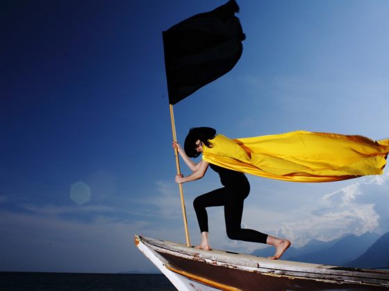 Eine schlanke junge Frau, die ein enges schwarzes Trikot, einen schwarzen, großen Hut und einen wehenden, leuchtend gelben Umhang trägt, steht auf dem Bug eines Bootes und schwenkt eine große schwarze Fahne. Sie fährt über ruhiges Wasser gegen einen blauen Himmel.