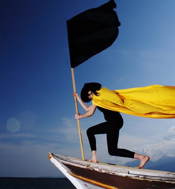 Eine schlanke junge Frau, die ein enges schwarzes Trikot, einen schwarzen, großen Hut und einen wehenden, leuchtend gelben Umhang trägt, steht auf dem Bug eines Bootes und schwenkt eine große schwarze Fahne. Sie fährt über ruhiges Wasser gegen einen blauen Himmel.