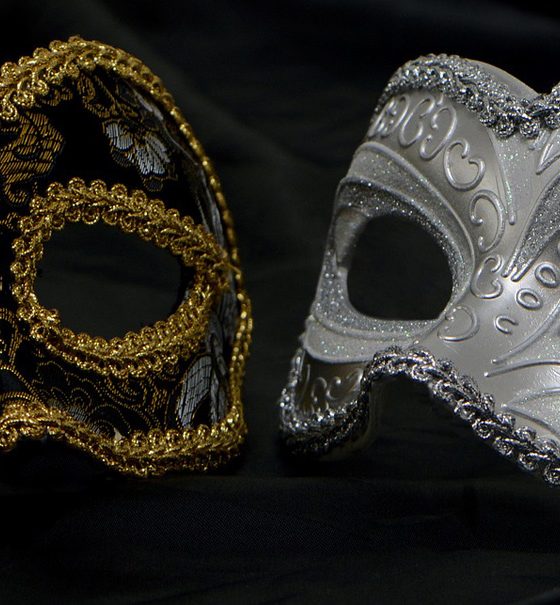 Zwei halbe venezianische Augenmasken liegen sich auf schwarzem Samt so gegenüber, dass sie ein Ganzes bilden. Sie sind beide mit Ornamenten bestickt, die eine Maske ist silbern, die andere schwarz und mit goldenen Säumen verziert.