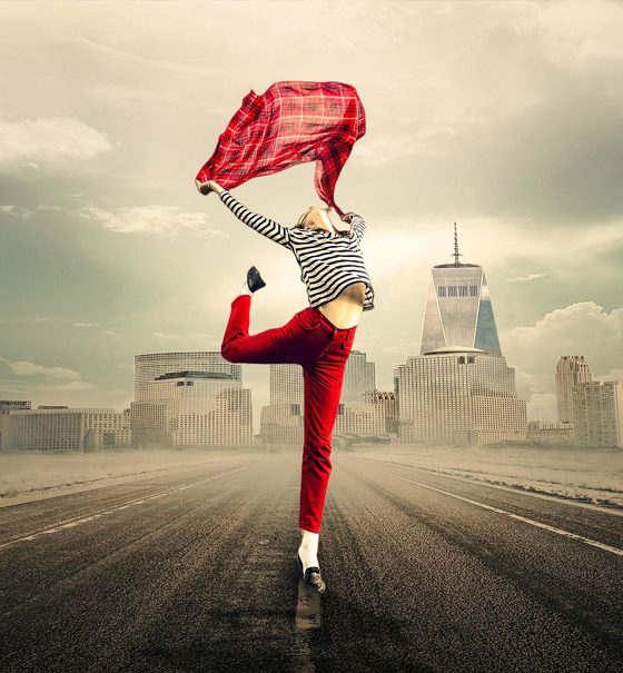 Eine schlanke junge Frau in einer roten Hose und einem blauweiß gestreiften Oberteil tanzt ekstatisch auf einer sehr breiten Straße vor einer Großstadt-Silhouette. Sie reckt das rechte Bein nach hinten hoch in die Luft und schwenkt ein rot kariertes Tuch mit beiden Armen über ihrem Kopf.