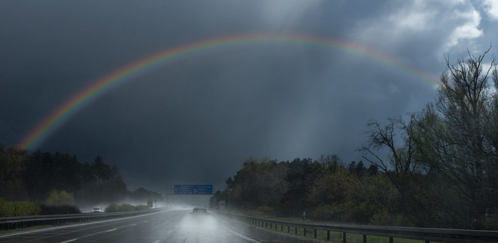 Ein Regenbogen überspannt eine breite Autobahn, der Himmel ist bedrohlich dunkelgrau.