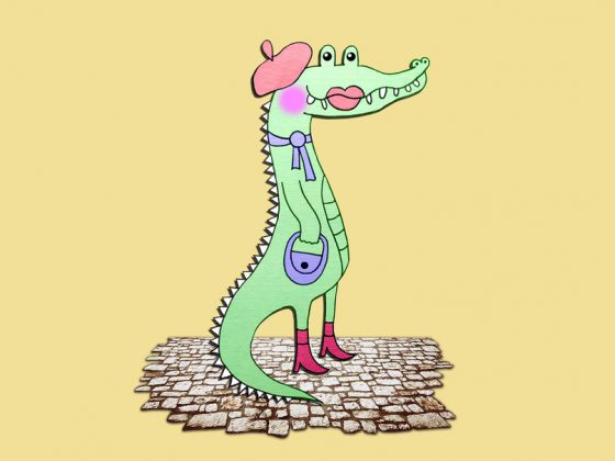 Auf einem zartgelben Hintergrund steht ein gezeichnetes hellgrünes Krokodil in Profilansicht auf den Hinterbeinen. Es trägt rote Stiefeletten an den Füßen, ein hellblaues Handtäschchen am Arm und eine hellblaue Schleife um den Hals, sowie eine rosa Baskenmütze auf dem Kopf. Geschminkt ist es mit einem rosa Bäckchen und auf dem Maul aufgemalten Lippen.