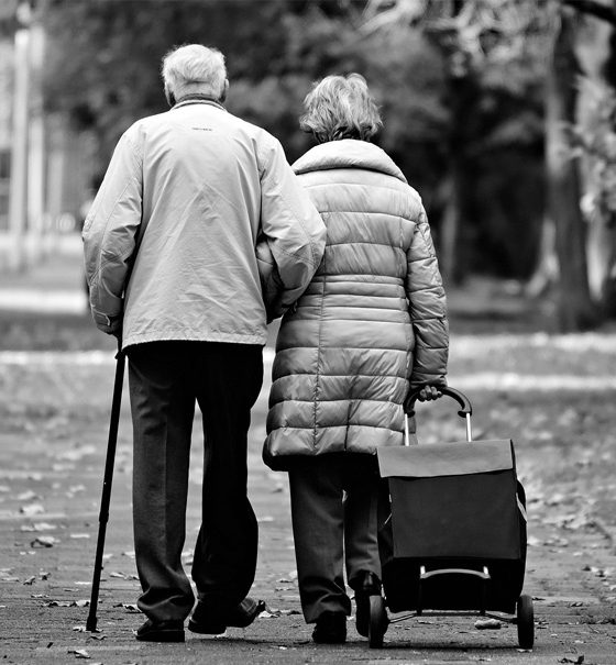 Die Schwarzweiß-Fotografie zeigt die Rückansicht eines alten Paares, das untergehakt eine Straße entlanggeht.