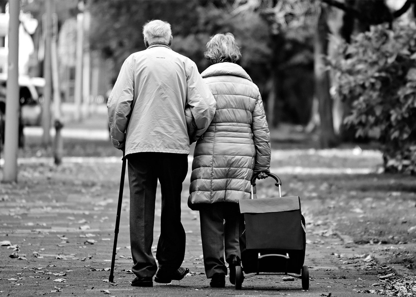 Die Schwarzweiß-Fotografie zeigt die Rückansicht eines alten Paares, das untergehakt eine Straße entlanggeht.